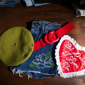Green beret, denim shorts with floral embroidery, red belt, and heart-shaped pillow with love message on wooden surface.
