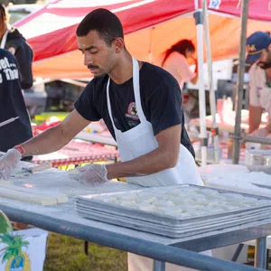 Man wearing apron preparing dough at outdoor food stall with trays of dough and tents in background