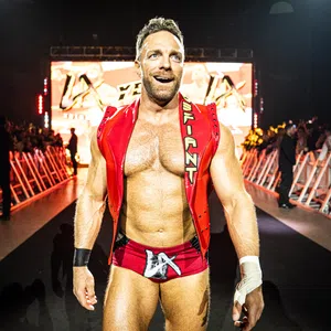 Muscular male wrestler in red trunks and vest walking confidently toward ring with audience and lights behind him