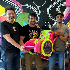Group of five people smiling indoors with colorful wall art, holding a bright pink and green custom wheelchair.