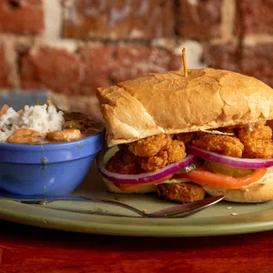 Crispy fried shrimp po' boy sandwich with lettuce, tomato, onion, served with a side of rice and beans in a blue bowl.
