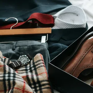 Flat lay of men's brown dress shoes in a box, plaid scarf, gray trousers, watch, and a white shirt on a dark suit jacket.