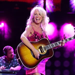 Female country singer in pink dress playing acoustic guitar on stage with vibrant purple lighting and smiling joyfully.