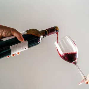 Red wine being poured from a bottle into a glass held by two hands against a plain background.