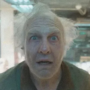 Older man with wild white hair and surprised expression looking directly at the camera indoors.