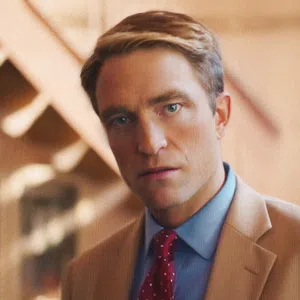 Man in a tan suit and red polka dot tie staring directly at the camera with a blurred warm background.