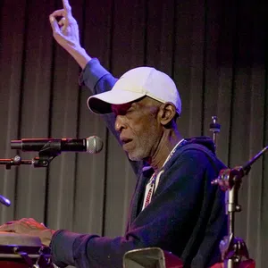 Elderly male musician playing congas and singing into a microphone with one hand raised on stage.