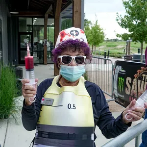 Person in nurse costume wearing sunglasses and mask holding two large syringes outside a building.