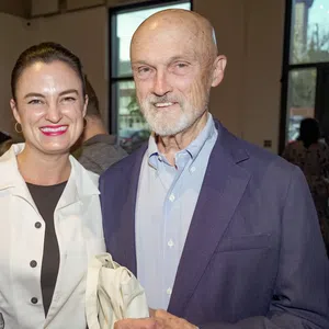 Smiling woman and elderly man standing indoors at a social event with large windows in the background.