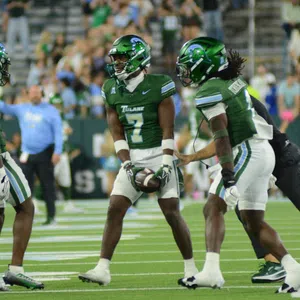 Tulane football players in green uniforms huddle on field during game with crowd in background