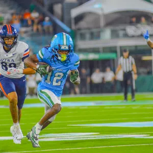 Football player in blue uniform running with ball as opponent in white uniform chases him on the field.