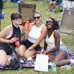 Three women wearing sunglasses enjoying drinks while sitting on a blanket at an outdoor event on grass.