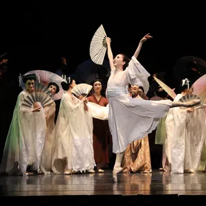 Ballet dancer in white costume performs with fans on stage with other dancers in traditional attire.