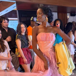 Model in pink gown holding yellow jacket walking past crowd of spectators at indoor event with wood paneling.