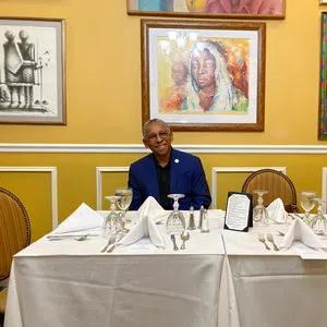 Man in blue suit sitting at white tablecloth-covered table set with wine glasses and silverware, framed art on yellow wall.