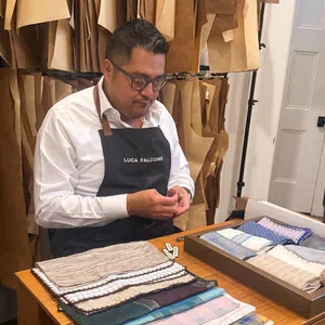 Tailor wearing Luca Falcone apron working with fabric samples in a workshop filled with hanging patterns.