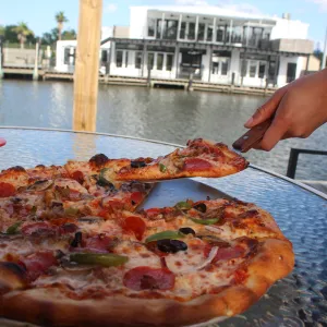 Person lifting a slice of pepperoni pizza from a glass table by the waterfront with a house in the background