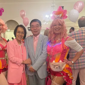 Two women in colorful circus costumes and pink wigs pose with a couple dressed in pastel pink and gray at a festive event.