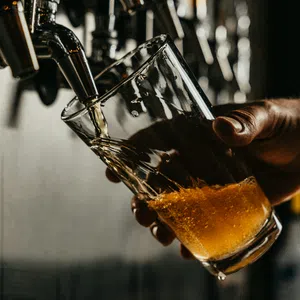 Hand pouring amber beer from tap into tilted glass held by hand in dimly lit bar setting.