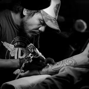 Black and white photo of a tattoo artist focused on inking a geometric tattoo on a client's forearm in dim light.