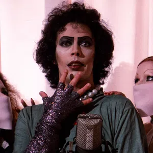 Actor in iconic glam rock makeup and costume with two masked fans on either side against a pale curtain.