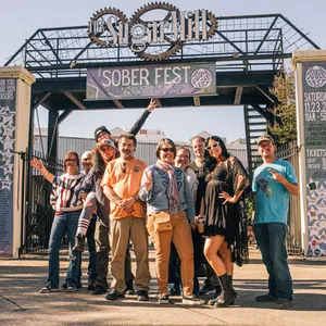 Group of people posing and smiling under Sober Fest banner at outdoor event entrance on sunny day