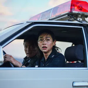 Female police officer and male colleague in a police car with red siren light, looking concerned through open window.