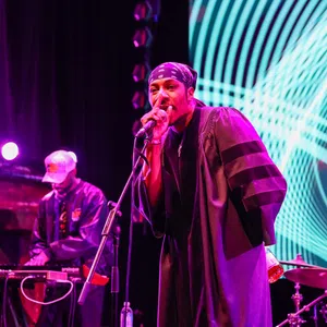 Hip-hop artist performing on stage with microphone and DJ in the background under vibrant purple and blue lights.
