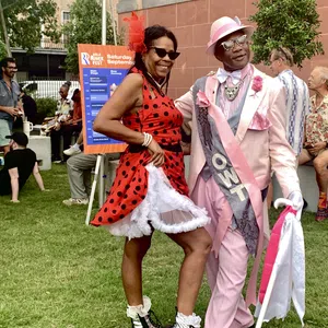 Couple dressed in vibrant retro outfits enjoying an outdoor festival on a sunny day with people relaxing nearby