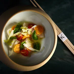 Elegant bowl of fresh vegetable salad with cherry tomatoes, greens, and chopsticks on dark surface.