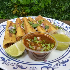Plate of golden taquitos garnished with cilantro and onions, served with lime wedges and two dipping sauces.