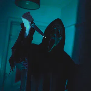 Person wearing a ghost face mask and dark robe holding a raised knife in a dimly lit room.
