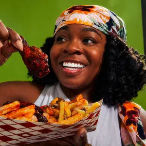 Smiling woman holding a saucy chicken wing and a tray of seasoned fries against a green background.