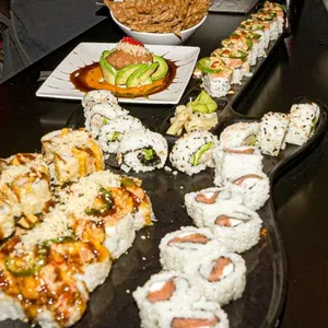 Variety of sushi rolls arranged on a black table with sauces, avocado, and garnishes in a restaurant setting.