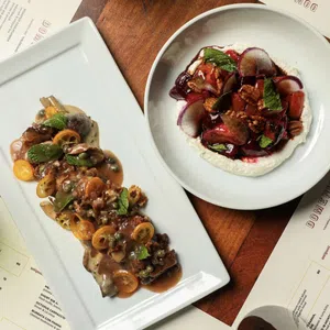 Gourmet dishes with sauces and herbs served on white plates surrounded by menus and wine glasses on wooden table.