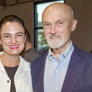 Smiling woman and older man standing indoors near a window in a casual social setting.
