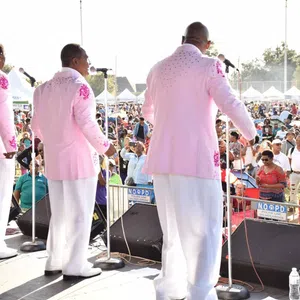 Five male singers in pink jackets performing on outdoor stage before large audience at daytime event.