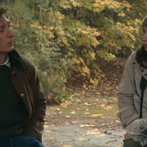 Man and woman dressed in autumn clothing sit outdoors surrounded by fall foliage during a conversation.
