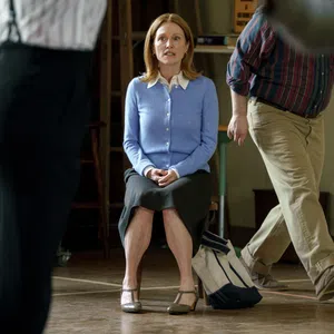 Woman in blue cardigan and black skirt sitting on a chair surrounded by blurred walking people indoors