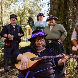 Group of six people in medieval and pirate costumes posing outdoors with a lute player in front under a large tree.