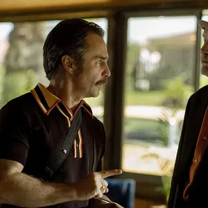 Two men in conversation, one wearing a hat and suit, the other in a black shirt with orange collar details indoors near windows.