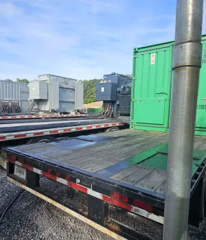 Flatbed trailers with industrial equipment and green shipping containers parked outdoors on gravel.