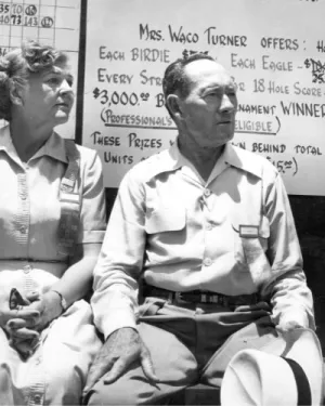 Black and white photo of a man and woman sitting in front of a handwritten golf prize board.