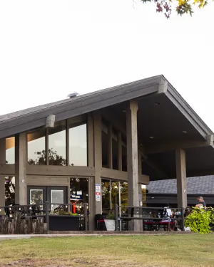 Modern rustic cafe with large glass windows and outdoor seating under a sloped roof surrounded by greenery.