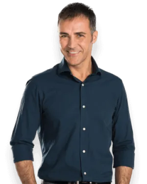 Smiling man in a dark blue button-up shirt and gray pants with hands in pockets against a black background