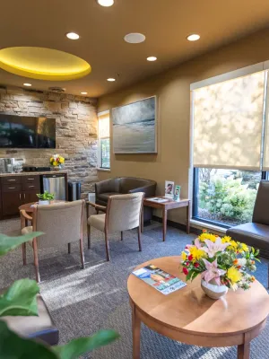 Bright, cozy waiting room with beige chairs, stone accent wall, large windows with blinds, and fresh floral centerpiece.