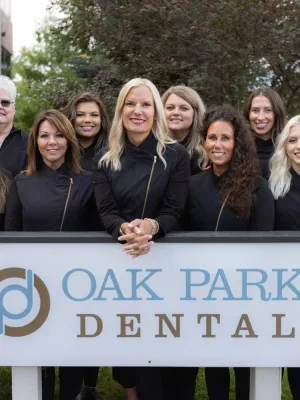Dental team of Oak Park Dental posing outdoors behind a sign with logo and name in matching black outfits.