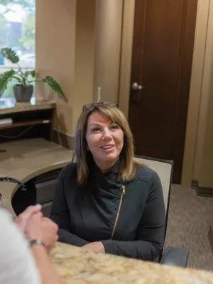 Smiling receptionist sitting at a desk talking to a visitor in a warmly lit office reception area.