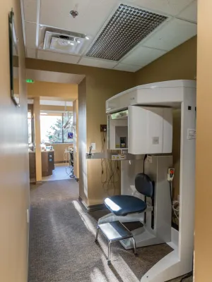 Dental X-ray machine with chair inside a beige-walled clinic hallway with natural light coming from windows.