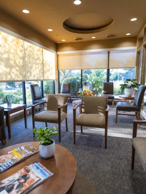Sunlit waiting room with chairs, a couch, magazines on a wooden table, and large windows with blinds.